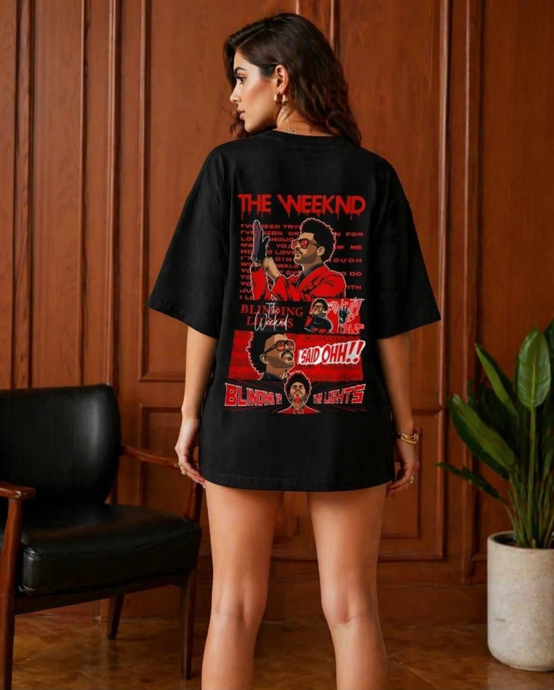 Person wearing a black t-shirt with graphic design and text, standing in a room with wooden paneling and a plant.