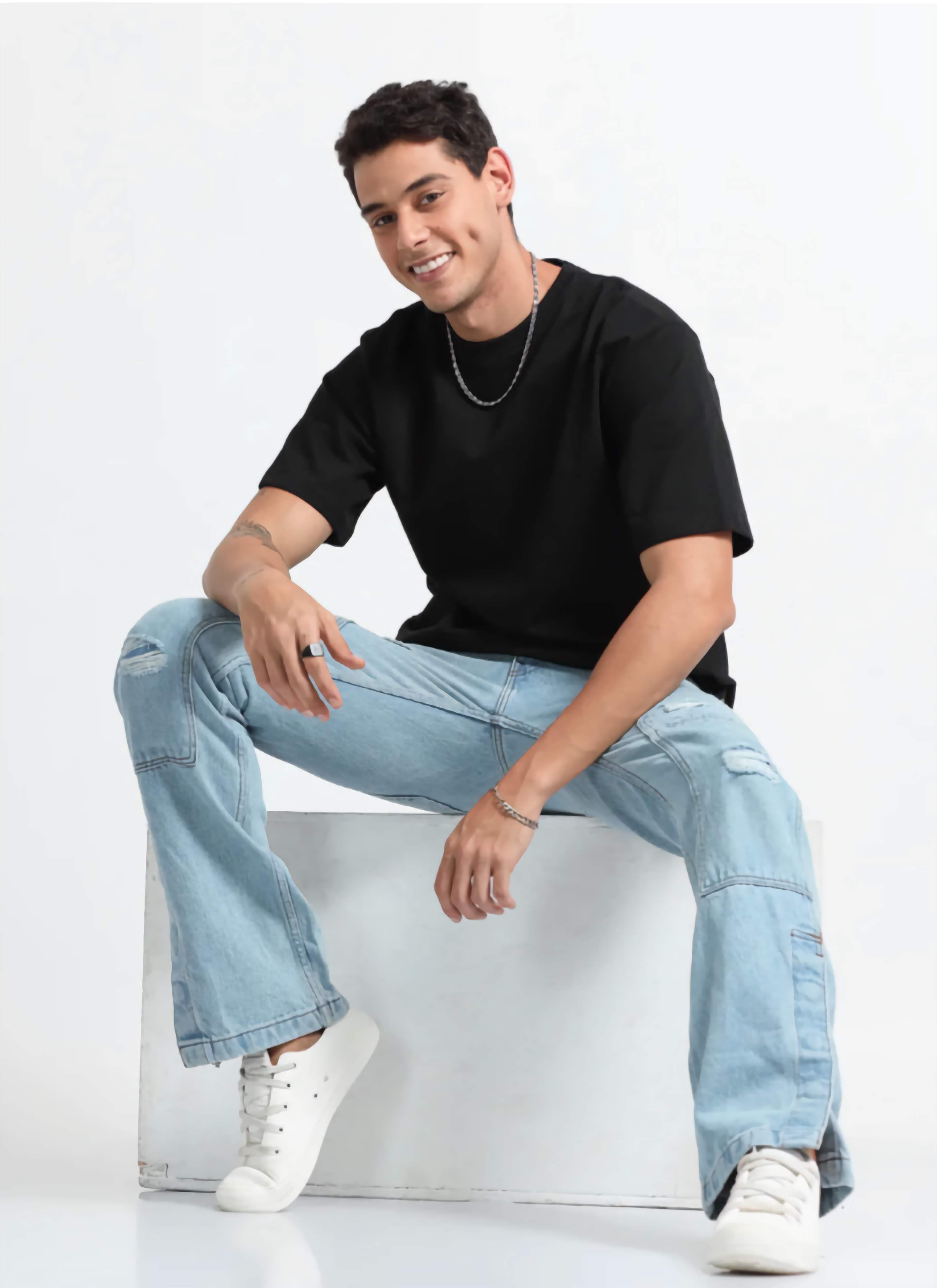 Man wearing a black t-shirt, light blue jeans, and white sneakers sitting on a white stool against a white background.