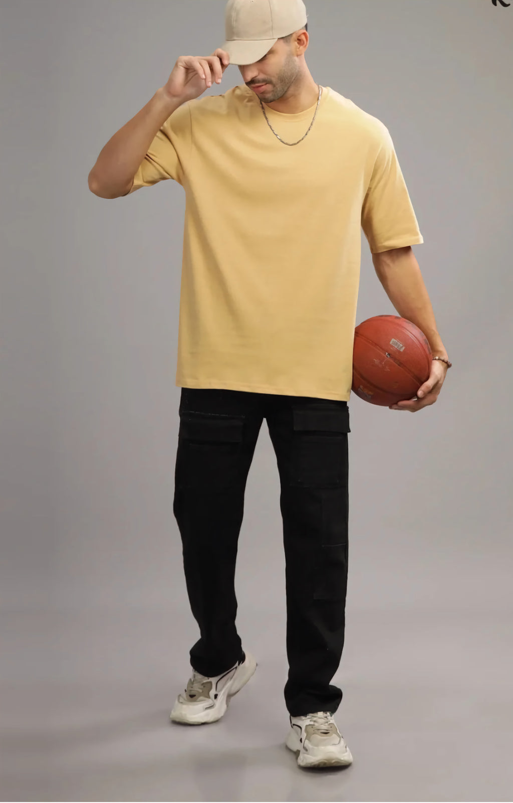 Man wearing a yellow t-shirt, black pants, and a beige cap, holding a basketball on a gray background