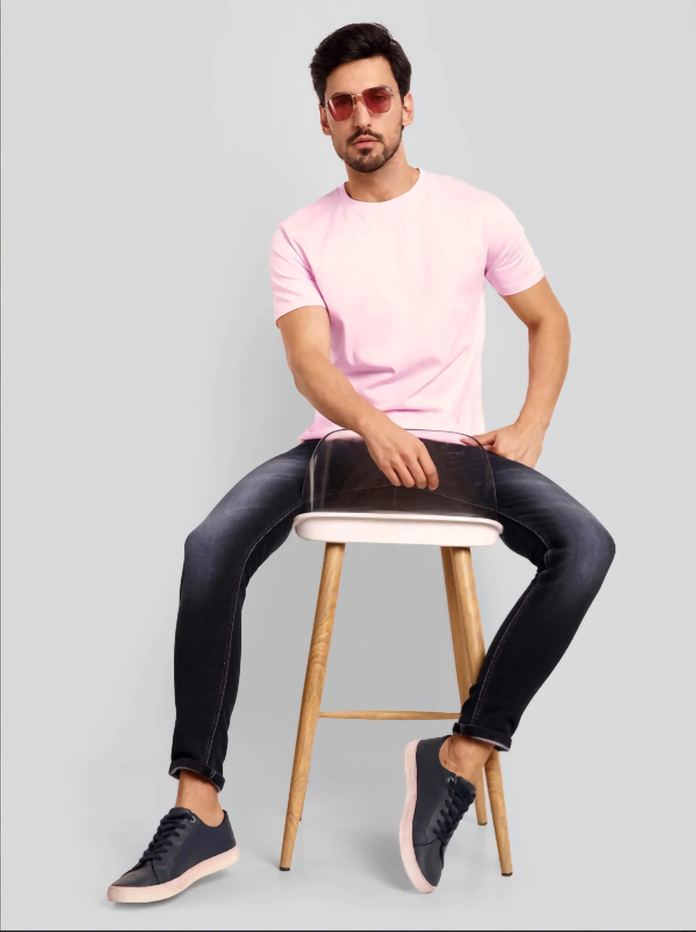 Man sitting on a stool wearing a pink t-shirt and black jeans against a gray background