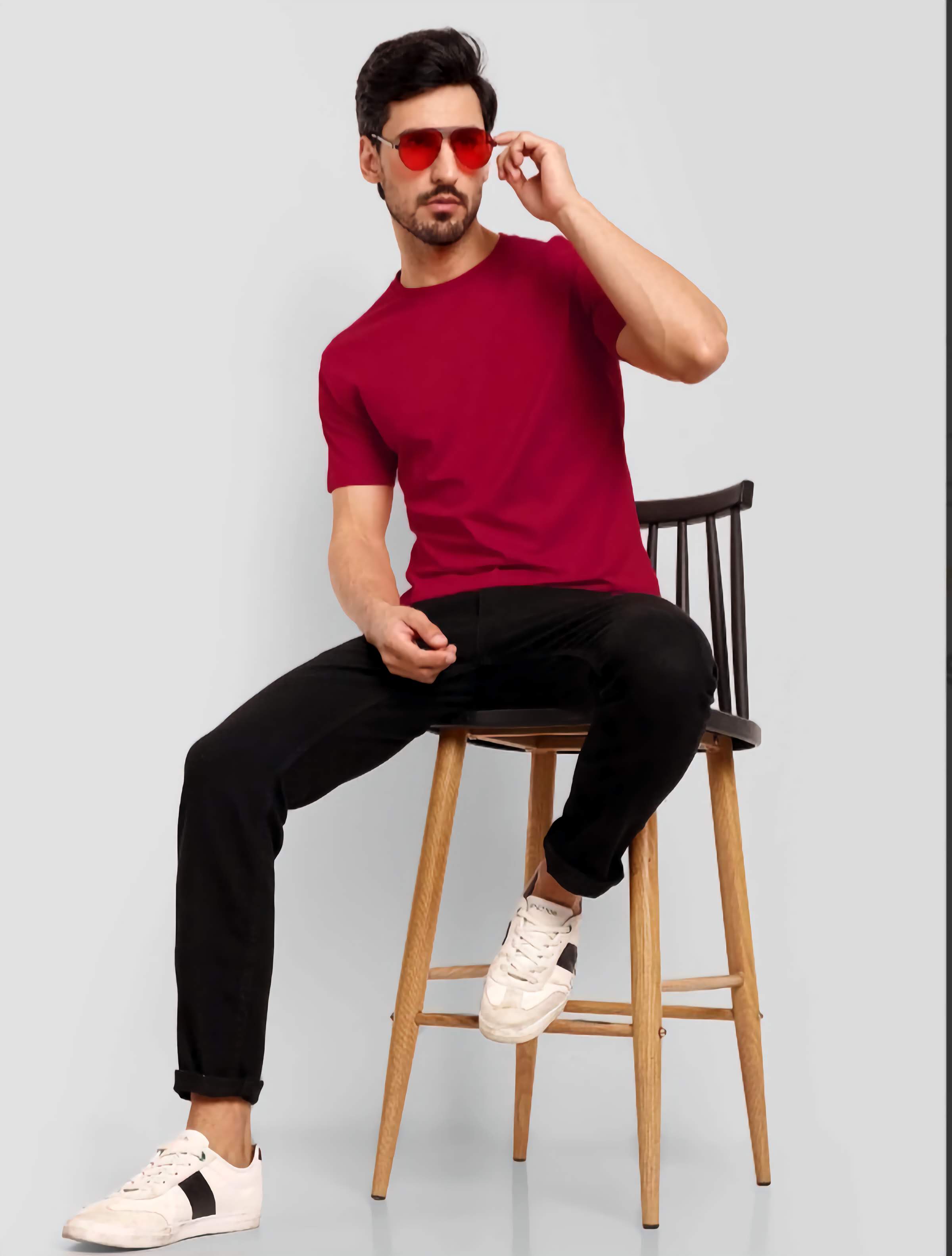 Man wearing a red shirt, black pants, and white sneakers sitting on a wooden stool against a light gray background.